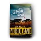 Text: Die Küste vergibt nicht. Ida Munk. Nordland. Kriminalroman.
Häuser in Dünenlandschaft, eine Flagge weht im Wind.