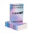 Buchcover: "Bernhard Aichner", "Yoko", "Thriller". Mehrere Bücher gestapelt, Design in Blau- und Pinktönen.