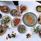 Ein Tisch mit einem festlichen Gedeck: Lachsfilet, Tomatensalat, Brotkorb, Käse, Blumen und Besteck auf weißem Tischtuch.
