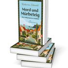 Ein Buchstapel zeigt "Mord und Mürbeteig" von Katharina Schendel. Das Cover hat eine Illustration eines Dorfes.