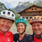 "Kronen Zeitung" steht auf roten Radtrikots. Drei Personen in Fahrradhelmen lächeln, dahinter eine Berghütte und grüne Berge.