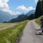Eine schmale Straße schlängelt sich durch grüne Wiesen, zwei Radfahrer fahren, Berge und ein Wald im Hintergrund.