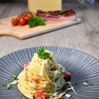 Ein Teller Spaghetti mit Käse, Speck, Tomaten und Petersilie; im Hintergrund Käse, Speck und Tomaten auf Holzbrett.