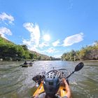 Ein Paddel mit "AQUA-BOUND" Text wird gehalten. Eine Person in einem gelben Kajak fährt auf einem Fluss bei sonnigem Wetter.