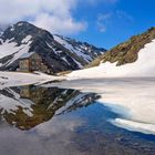Ein steinernes Gebäude spiegelt sich in einem klaren Bergsee, umgeben von schneebedeckten Bergen und blauem Himmel.