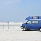 Ein blauer Campervan steht am Strand. Menschen und das Meer sind im Hintergrund zu sehen.