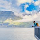 Eine Frau mit gelbem Rucksack lehnt an einem Holzbalkon vor einer dramatischen Berglandschaft mit Wolken.