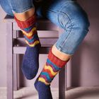 Keine sichtbaren Texte. Eine Person sitzt auf einem Stuhl und trägt bunte Stricksocken und blaue Jeans.