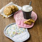 Tasse mit Schaum und Löffel auf rosa Untersetzer, daneben Kekse und ein floraler Untersetzer auf Holz.