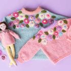 Zwei gestrickte Pullover, einer blau, einer rosa, mit Blumenmuster. Daneben liegt eine Puppe in rosa Kleidung.