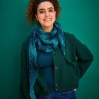 Eine Person lächelt vor einem grünen Hintergrund, trägt eine dunkle Jacke, blauen Pullover und Schal, mit lockigem Haar.