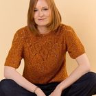 Eine lächelnde Person mit braunem Haar trägt einen orange-gemusterten Pullover und sitzt entspannt mit gekreuzten Beinen.