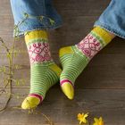 Grüne Socken mit Streifen und rosa Mustern, getragen von jemandem in Jeans. Daneben sind gelbe Blumen auf Holzboden.