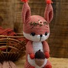 Amigurumi-Eichhörnchen mit Blumenkranz hält eine Eichel, sitzt vor einem Korb auf Holzuntergrund.