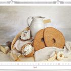 April. Eine rustikale Szene mit zwei großen Broten, Käse, einem Krug und Knoblauch, auf weißem Tuch arrangiert.