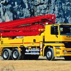 Text: Putzmeister, Elefant, Betonpumpe, 3331. Gelber Betonmischer-Lkw mit rotem Aufbau vor Felswand, Arbeiter daneben.