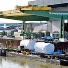 Schwergutkran mit "Schwergutzentrum KÜBLER SPEDITION". Lastschiff mit großen, verpackten Gütern am Wasser.