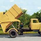 Roter Etikettentext auf einer großen Militärmaschine. Ein gelber Lastwagen mit geneigter Ladefläche, umgeben von Bäumen.