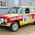 Rallyeauto mit Aufschriften „LUCKY STRIKE“, „NORTTI“, „61“. Rot-gelbes Design, parkend vor einem grauen Gebäude.