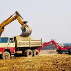 Zwei Lkws mit "Richter", ein gelber Bagger lädt Sand. Baustelle mit Wald im Hintergrund.