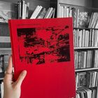 "CHRIS DORLAND: FUTURE RUINS" steht auf einem roten Buch mit schwarzem abstraktem Design. Eine Hand hält es vor einem Bücherregal.