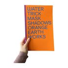 WATER TRICK MASK SHADOWS ORANGE EARTH WORKS. Orange Buchcover, lila Rücken, gehalten von hand in lila Ärmel.