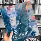 „HUNDERT, Ruprecht von Kaufmann, Hirmer“ auf einem farbenfrohen Buchcover mit abstrakter Illustration. Im Hintergrund ein Bücherregal.