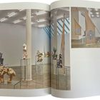 Seite 136, 137: Galerie mit Skulpturen auf Glasvitrinen, großer Stoffbanner mit Spiralmuster. Helle, moderne Umgebung.