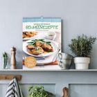 Ein Regal mit Pflanzen, einem Küchenutensil und einem Kalender. Der Kalender trägt den Titel "GENUSSREZEPTE 2027".