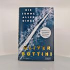 "Die Summe aller Dinge" von Oliver Bottini. Ein Luftbild eines Bootes im Meer, mit goldener Schrift auf dunkelblauem Hintergrund.