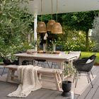 Keine Texte sichtbar. Ein eleganter, moderner Garten mit Holztisch, Bänken, schwarzen Stühlen und hängenden Rattanlampen.