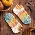 Strick-Socken in sanften Regenbogenfarben, Garnknäuel, Schere und Stricknadeln auf einem Holztisch arrangiert.