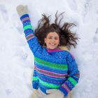 Eine lächelnde Person liegt im Schnee, trägt einen bunten Pullover und beige Handschuhe. Ihr Haar ist ausgebreitet.