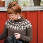 Eine Frau mit Brille strickt; sie trägt einen braunen Pullover mit weißen Mustern und sitzt vor einer roten Holzwand.