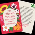 Eine Karte zeigt den Text: „Mit offenen Armen lässt es sich besser umarmen.“ Umgeben von bunten Blumenillustrationen.