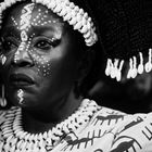 Ein Mensch trägt traditionelle Kleidung mit Tiermotiven, Perlen und Muscheln im Haar und weiße Gesichtsbemalung in geometrischen Mustern.