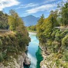 Kein Text vorhanden. Ein Fluss fließt durch eine Schlucht, umgeben von Bäumen, mit Bergen im Hintergrund unter einem blauen Himmel.
