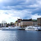 Aufschrift: "VINDHEM". Weiße Boote am Ufer, historische Gebäude im Hintergrund, darunter ein Segelschiff. Wolkiger Himmel.