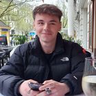 Junger Mann in schwarzer Jacke mit "The North Face"-Logo, hält Handy. Draußen an einem Tisch, daneben Glas mit weißem Getränk.