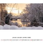 Januar. Ein winterliches Flussufer mit schneebedeckten Bäumen und Brücke im Hintergrund.