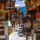 Schild: "Café Restaurant Crêperie La Claire Fontaine". Enge, gepflasterte Gasse mit traditionellen Holzhäusern und bunten Blumen.