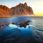 Kein Text vorhanden. Eine Berglandschaft spiegelt sich im ruhigen Wasser vor einem klaren Himmel beim Sonnenaufgang.