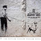 "THIS WALL IS A DESIGNATED GRAFFITI AREA." Ein Polizist und ein Pudel in Schwarz-Weiß sind an eine Ziegelwand gesprüht.