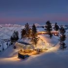 Ein Skilift beleuchtet im Schnee, umgeben von Baumgruppen, mit einem Halbmond am Abendhimmel und bergiger Landschaft im Hintergrund.