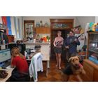 Eine Frau sitzt am Computer, während dahinter eine lächelnde Frau und ein Mann Gewehre halten. Ein Hund steht vorne.