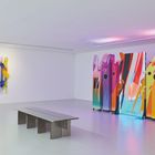 Eine moderne Kunstinstallation in einem hellen Raum. Links bunte Wandobjekte, rechts farbenfrohe, abstrakte Tafeln.