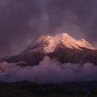 Ein schneebedeckter Berggipfel ragt über dunklen Wolken hervor, in violettem Licht getaucht.