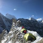 Zwei grüne Zelte stehen auf einem schneebedeckten Berggrat vor imposanten, verschneiten Gipfeln und strahlendem Sonnenschein.