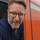 Ich kann den Text nicht erkennen. Ein Mann mit Brille lehnt aus einem orangefarbenen Zugfenster.