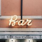 Schriftzug "Bar" in Neon, darunter Text "BAR San Calisto" auf grauem Schild, beige Fassade mit Lampe.